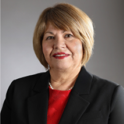 Photograph of Emma Torres. She is smiling, wearing a pearl necklace, has short blonde hair, and is wearing a red colored shirt and a black colored jacket.