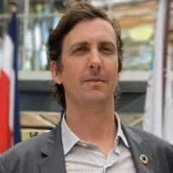 Photograph of Jason Glaser. He is in an outdoor urban space in front of a buildingand is wearing a light blue colored shirt and a slate grey colored jacket.